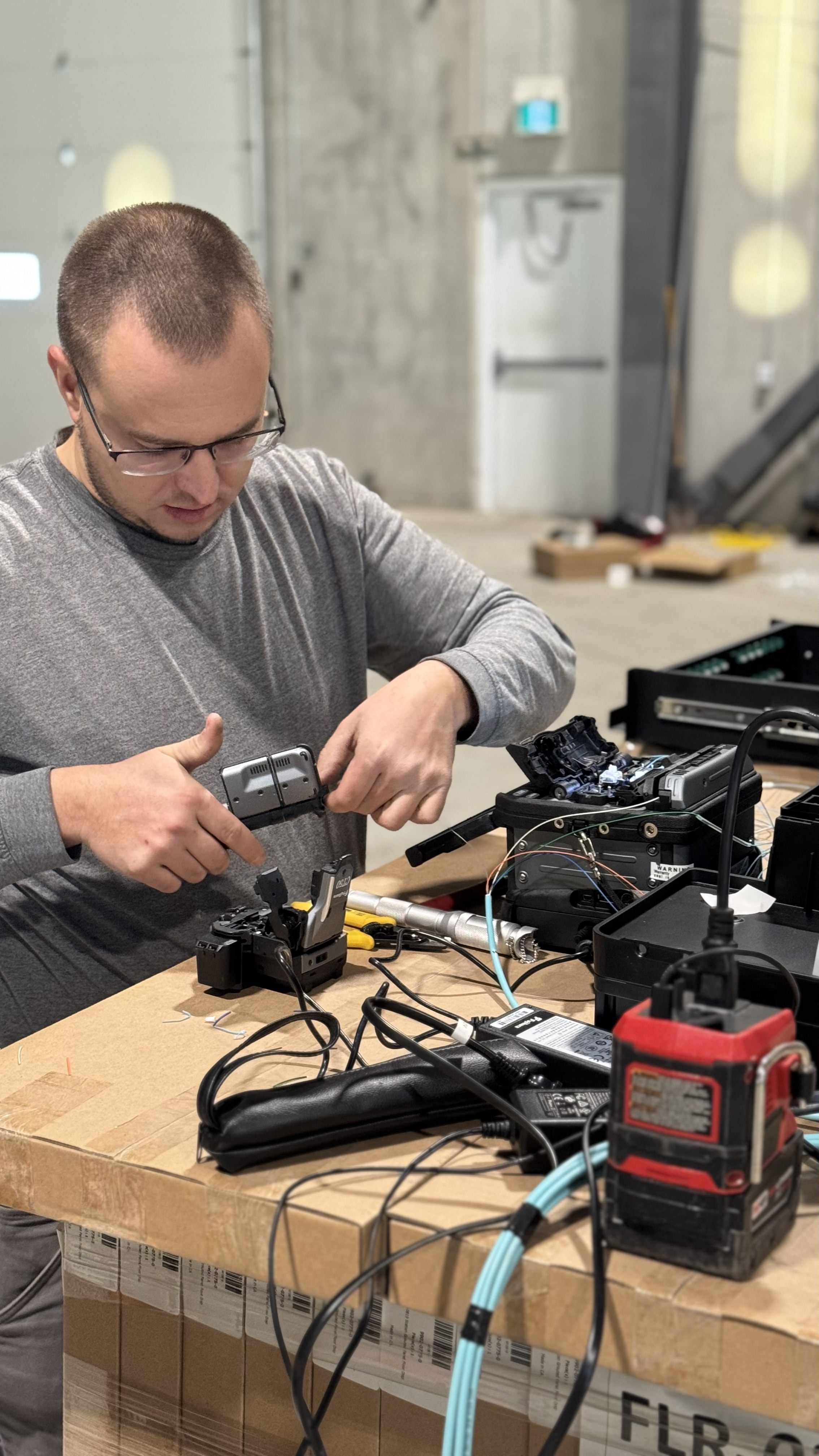 A man wearing glasses and a gray shirt is working on a table. He is focused on a piece of technical equipment with wires and cables, which he is holding with both hands. Other tools and equipment, including a gray device and a red power supply, are scattered across the brown cardboard table. The background shows a large, industrial-style room with a garage door and a white door with an exit sign above it.