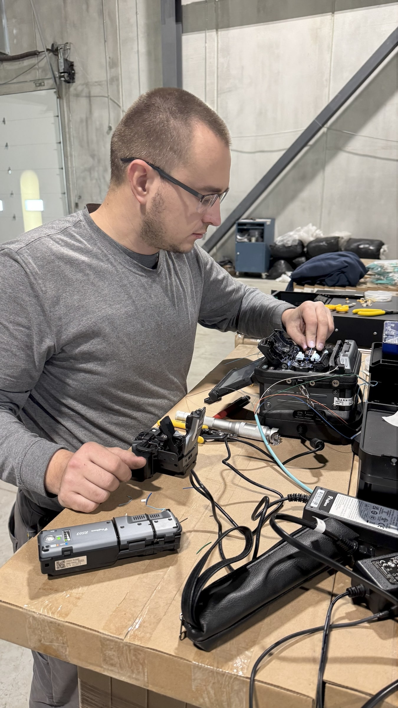 A close-up shot of a man wearing glasses and a gray shirt working on a table. He is focused on a piece of technical equipment with wires and cables. Other tools and equipment, including a gray device, are scattered across the brown cardboard table. The background shows a large, industrial-style room with a garage door.