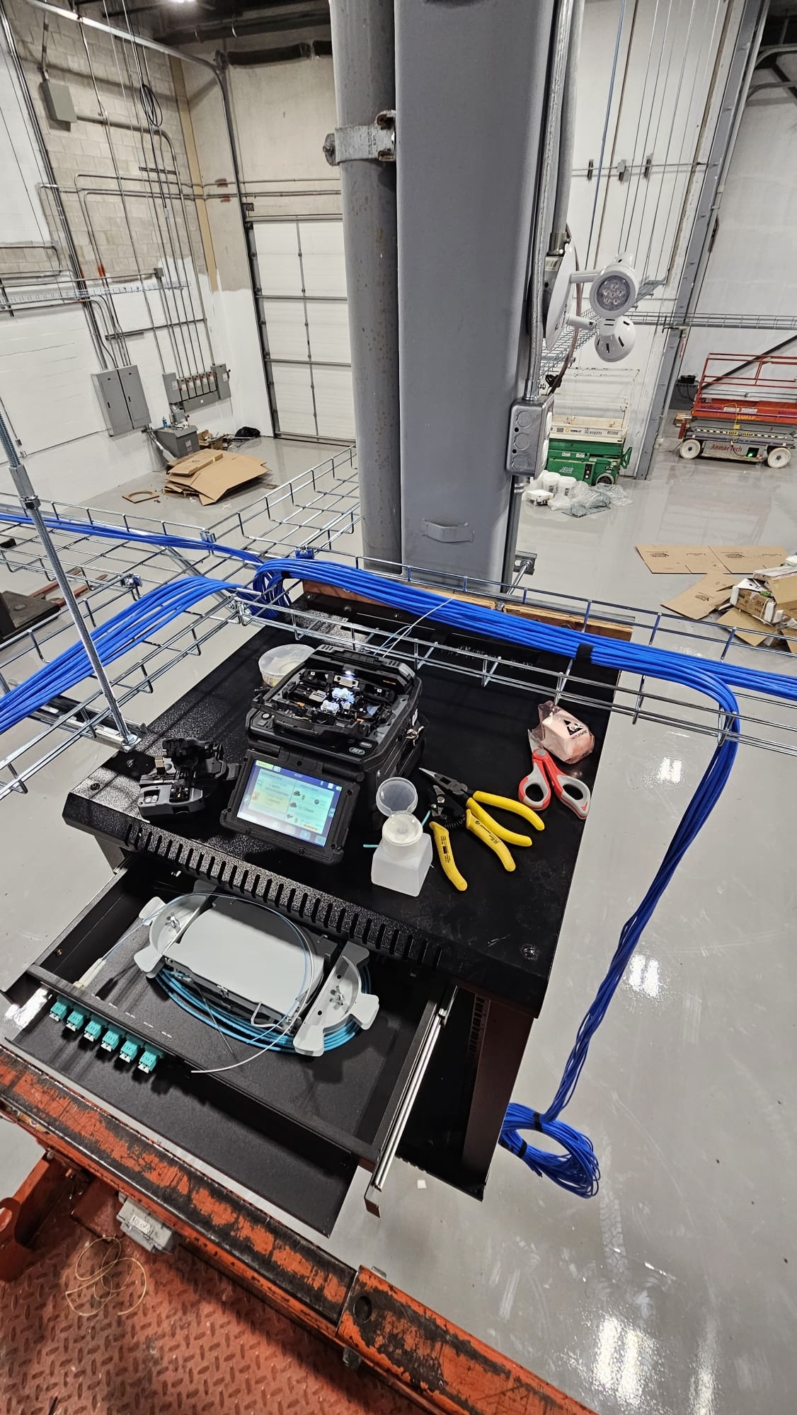 A top-down view of a **fiber optic cable** splicing and testing station set up on a work platform. The station includes a black **splicing machine** with a screen, a drawer holding a white **fiber splice tray** with a bundle of light blue cables, and various tools like yellow wire cutters and scissors. A bundle of blue network cables runs in a wire mesh cable tray on the left. In the background, a large garage door is visible, and the room has an industrial aesthetic with exposed pipes and ceiling structures.