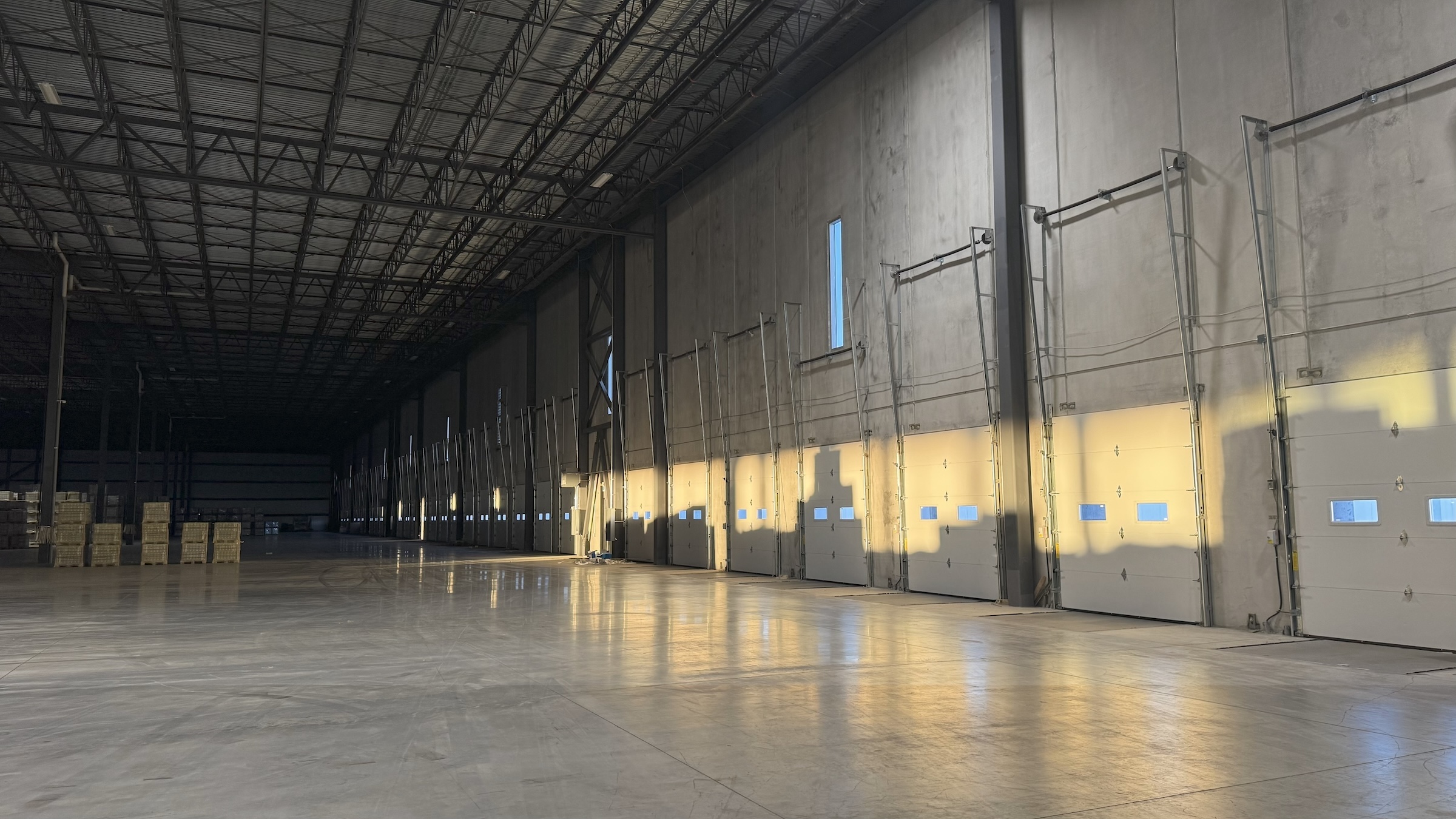 A wide, interior view of a large, empty warehouse or industrial building. The high ceiling is supported by a metal truss system. On the right, a series of closed white garage doors with small windows are illuminated by sunlight, which casts long shadows onto the polished concrete floor. Some boxes are stacked in the distance on the left.