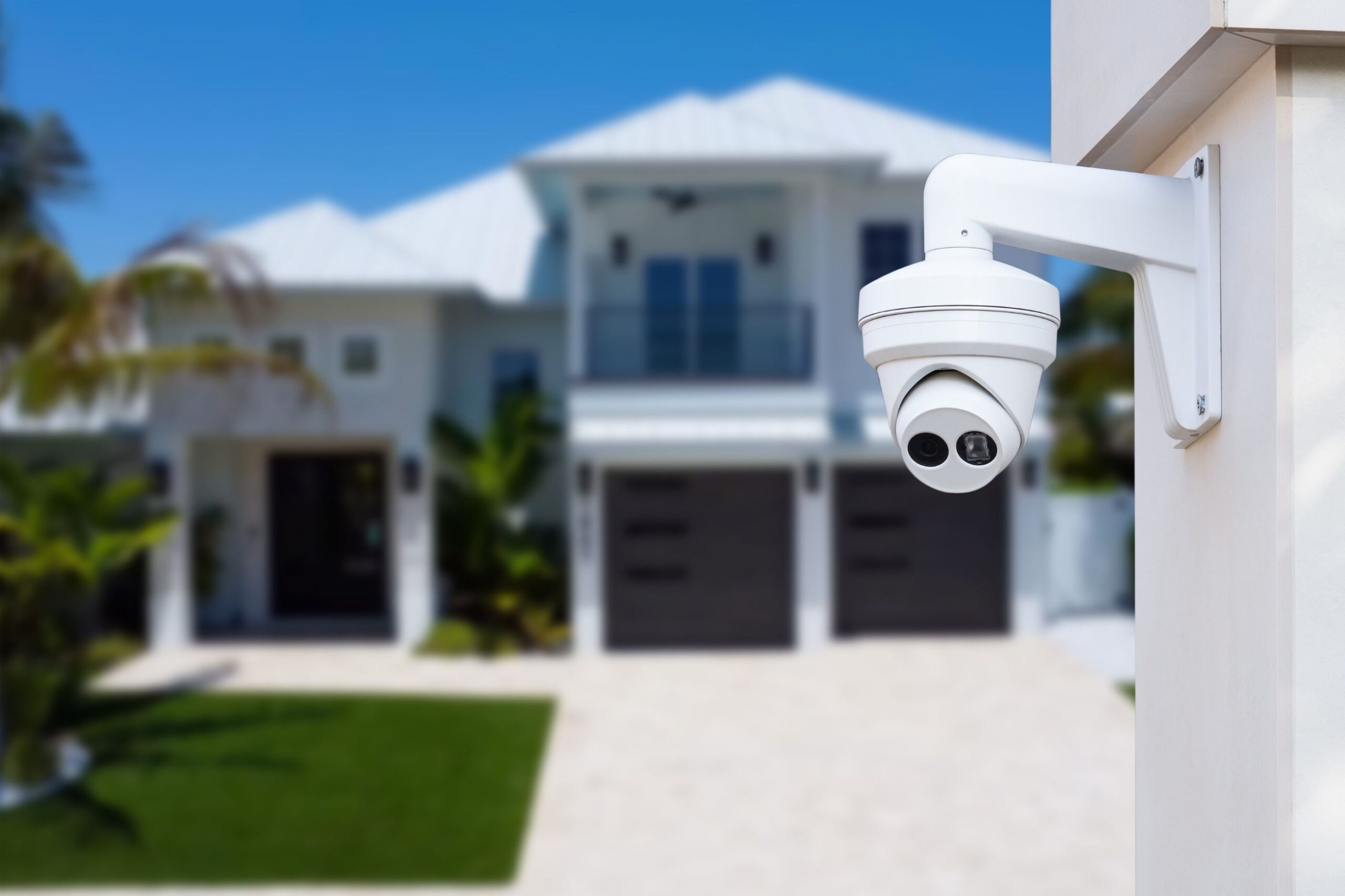 Close-up of a white dome security camera mounted on a pillar with a blurred modern two-story luxury home and palm trees in the background.