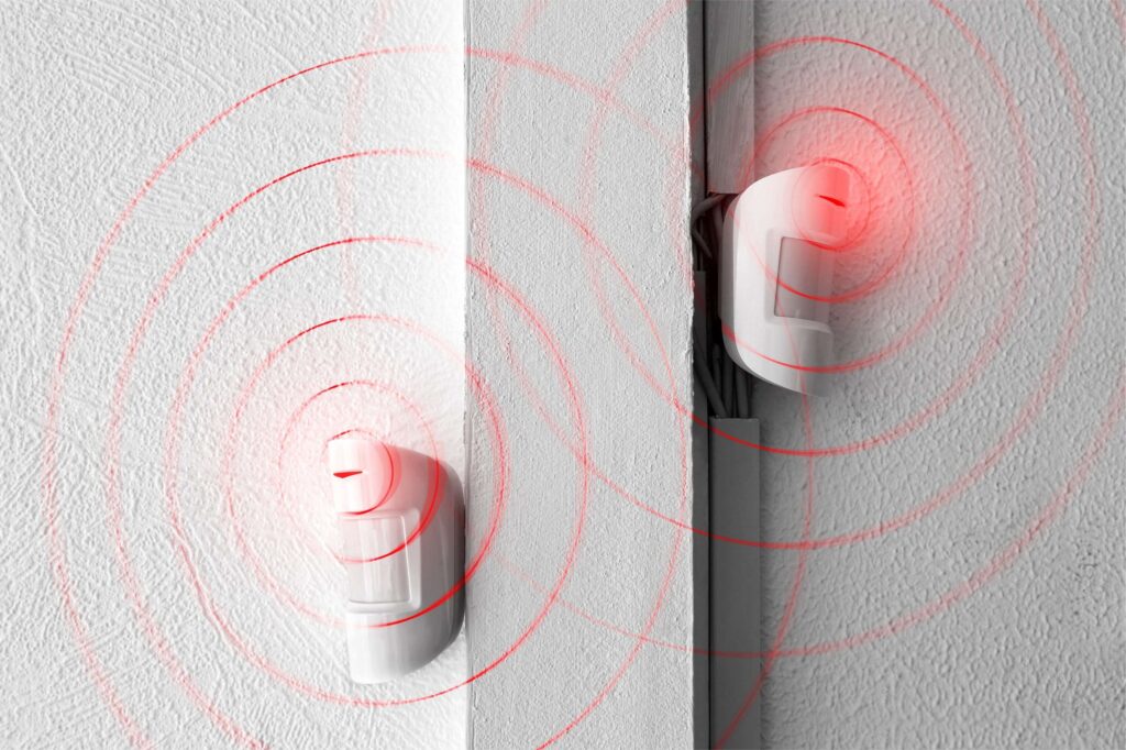 Two white motion detectors mounted on a white wall with red circular graphics representing infrared signal waves for intrusion detection.